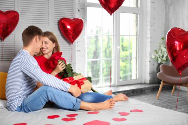 Kırmızı gül ve yatağa evde oturan hediye kutusu ile mutlu genç çift
