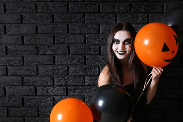 Beautiful woman with Halloween makeup and balloons standing near dark brick wall