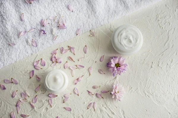 Jars with body cream, flower petals and soft towel on white table