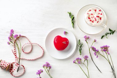Kalp şeklinde tatlı, fincan sıcak içecek ve beyaz ahşap zemin üzerinde çiçekler ile kompozisyon