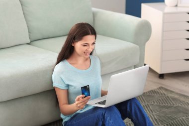 Genç bir kadın evde kredi kartı ve laptop ile online alışveriş yapıyor.