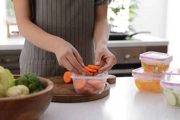 Woman putting fresh carrot slices into plastic container for freezing at table in kitchen