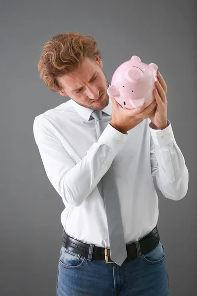 Young man tipping money from piggy bank on grey background