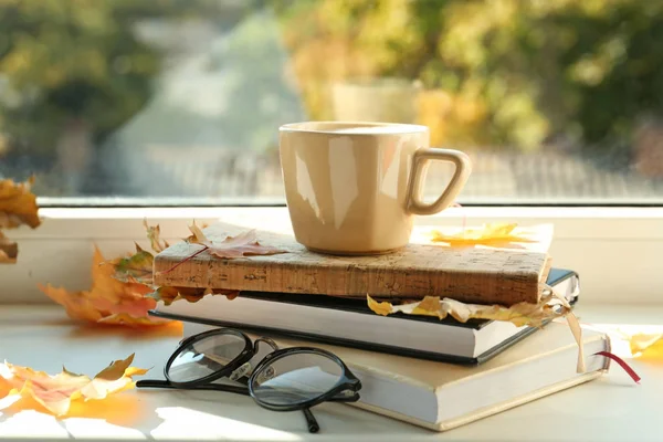 Cup of hot coffee with books and autumn leaves on windowsill