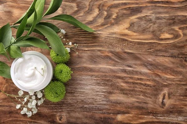 Jar with body cream and flowers on wooden table, top view