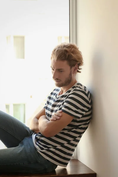 Depressed young man near window at home