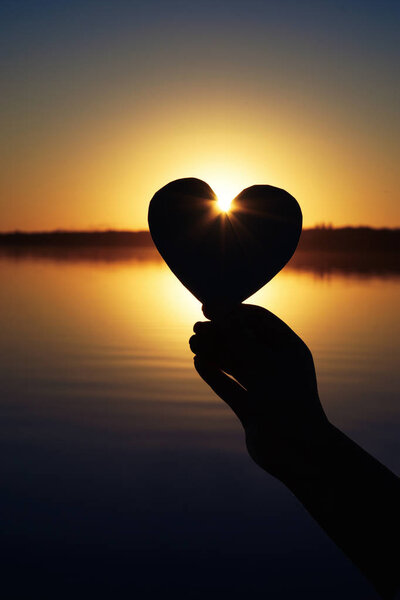 Hand of woman holding paper heart outdoors at sunset