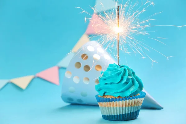 Tasty birthday cupcake with sparkler and party hat on color background