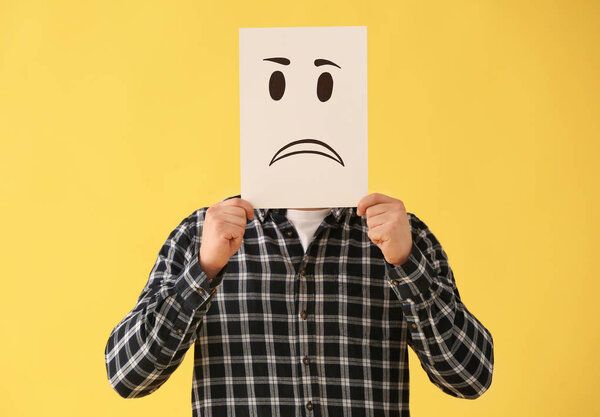 Человек, прячущий лицо за листом бумаги с нарисованным смайликом на цветном фоне
