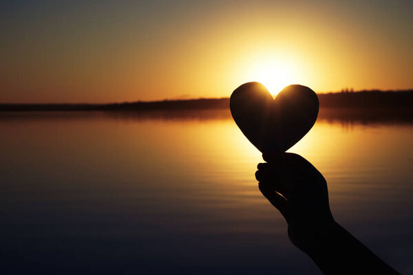 Hand of woman holding paper heart outdoors at sunset