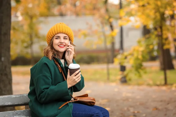 Beautiful young woman with coffee talking on mobile phone in autumn park