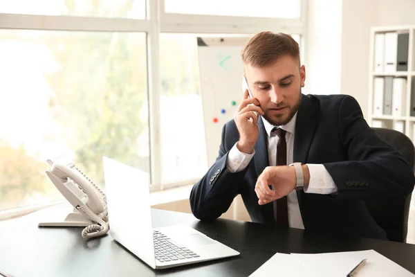 Businessman looking at watch while talking by phone in office