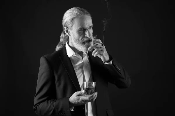 Black and white portrait of elderly businessman with glass of whiskey and cigar on dark background