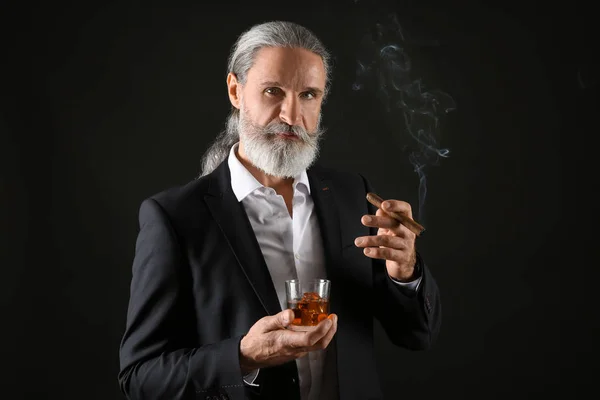 Elderly businessman with glass of whiskey and cigar on dark background