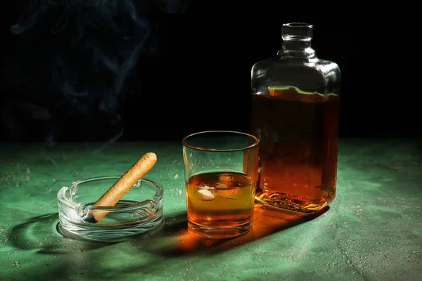 Bottle and glass of whiskey with cigar on color table against dark background