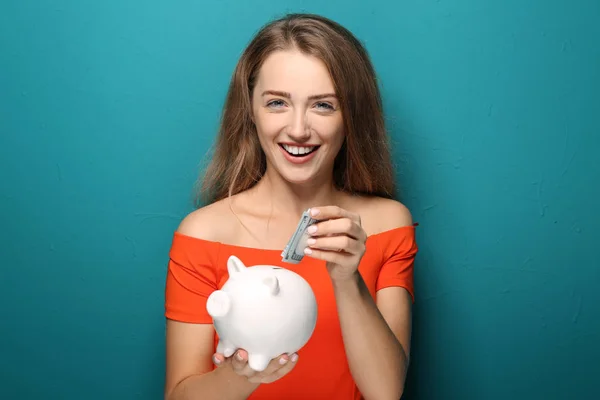 Young Woman Putting Banknote Piggy Bank Color Background — Stock Photo, Image