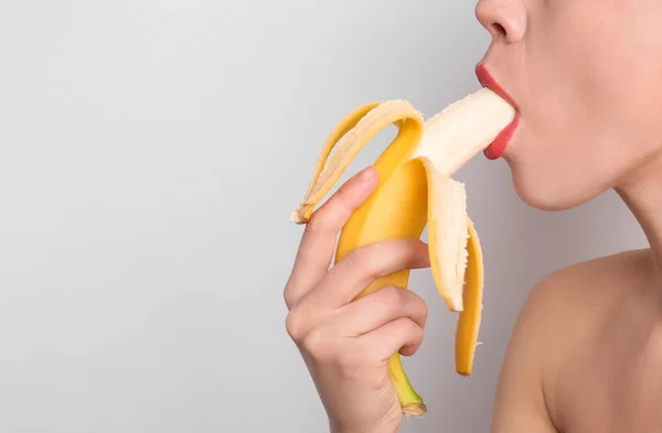 Sexy young woman eating banana on light background. Erotic concept