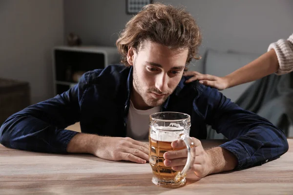 Man drinking beer at home and woman's hand on his shoulder. Alcoholism concept