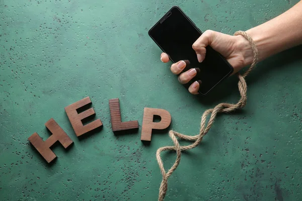 Woman with tied hand, mobile phone and word HELP on color background. Concept of addiction