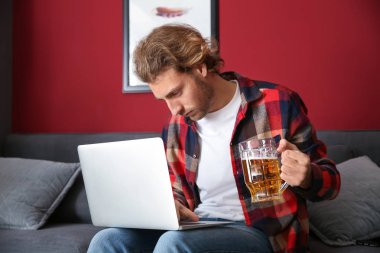 Genç adam dizüstü bilgisayar kullanıyor ve evde bira içiyor. Alkolizm kavramı