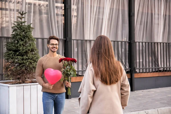 Young man with bouquet of roses and balloon waiting for his girlfriend outdoors