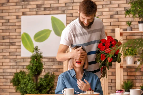 Man with bouquet of beautiful flowers covering his girlfriend\'s eyes in cafe