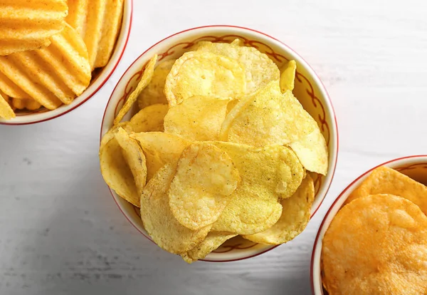 Bowls with tasty crispy potato chips on white wooden table