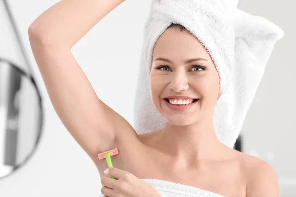 Beautiful young woman shaving armpits in bathroom