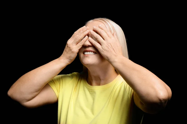 Portrait of beautiful mature woman covering eyes with hands on dark background