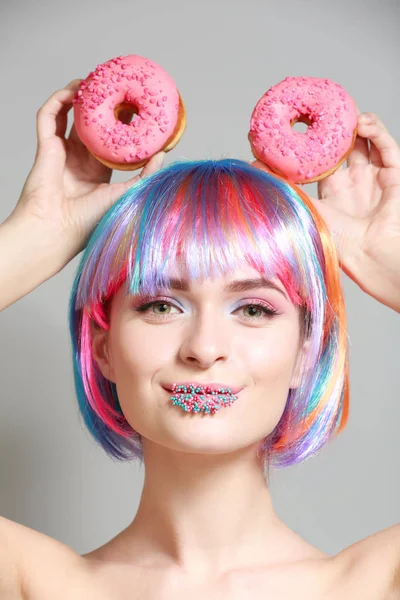 Beautiful young woman with unusual makeup and sweet donuts on grey background