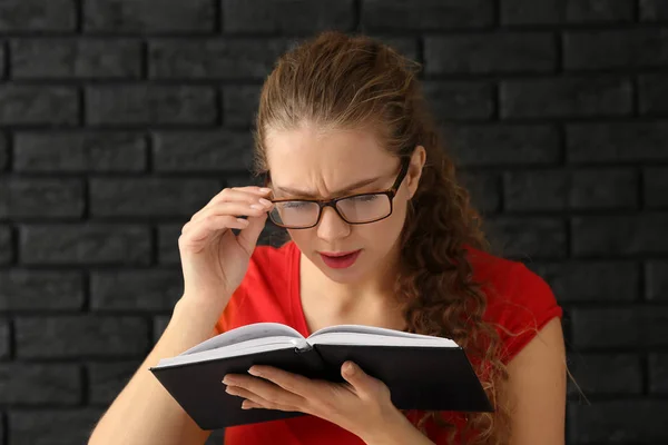 Young woman with bad sight reading book on dark brick background