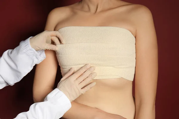 Doctor applying bandage on female chest after cosmetic surgery operation against color background