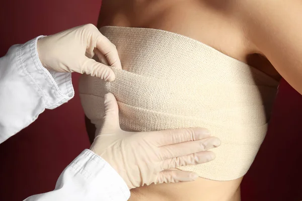 Doctor applying bandage on female chest after cosmetic surgery operation against color background