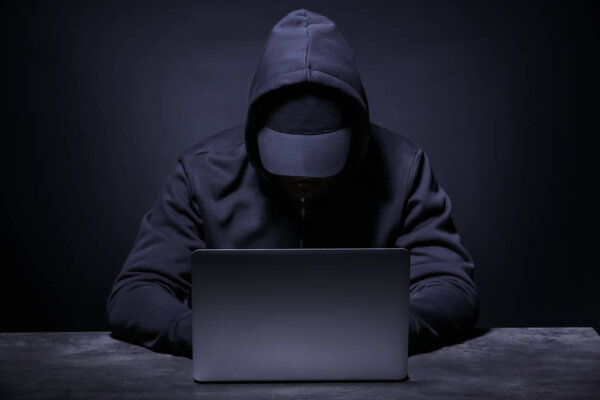 Professional hacker with laptop sitting at table on dark background
