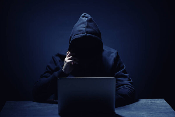 Professional hacker with laptop sitting at table on dark background