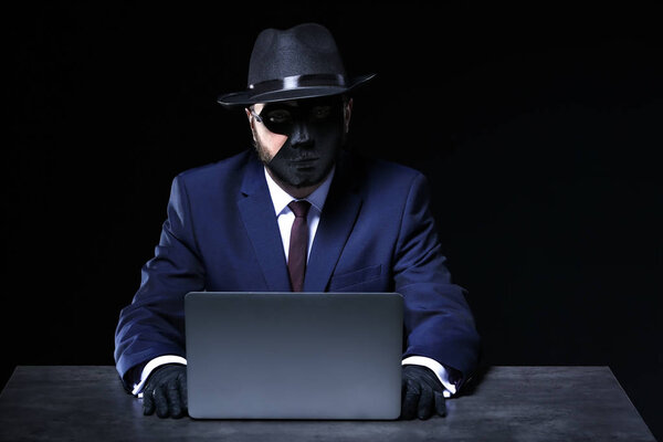 Professional hacker with laptop sitting at table on dark background