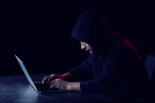 Professional hacker with laptop sitting at table on dark background