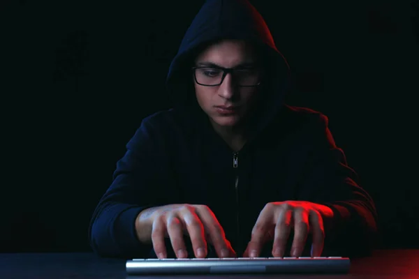 Professional hacker with computer keyboard sitting at table on dark background