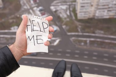 Depressed man holding paper sheet with text HELP ME on the edge of roof. Concept of suicide