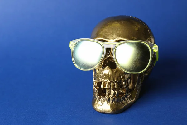 Golden human skull with sunglasses on color background