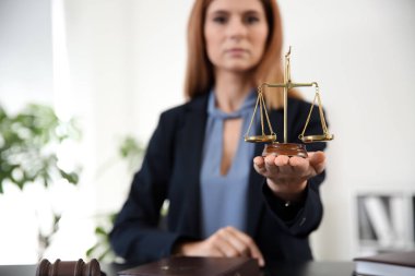 Beautiful female lawyer with scales of justice sitting at table in office clipart