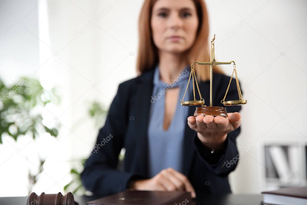 Beautiful female lawyer with scales of justice sitting at table in office