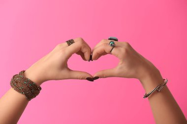 Young woman with stylish bijouterie making heart with her hands on color background