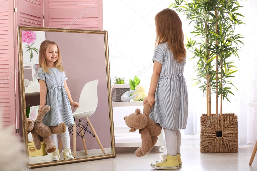 Cute little girl with teddy bear looking in mirror at home