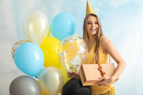 Beautiful young woman with balloons, party hat and gift on light background