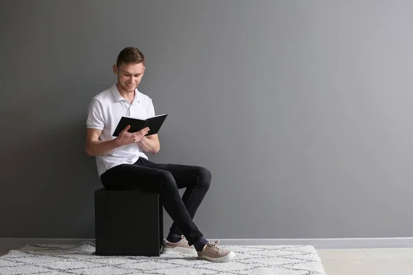 Bonito Jovem Homem Lendo Livro Enquanto Sentado Perto Parede Cinza — Fotografia de Stock