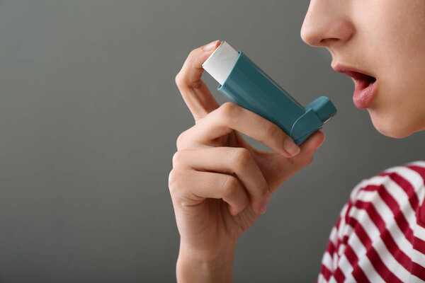 Молодая женщина, использующая ингалятор против астмы на сером фоне
