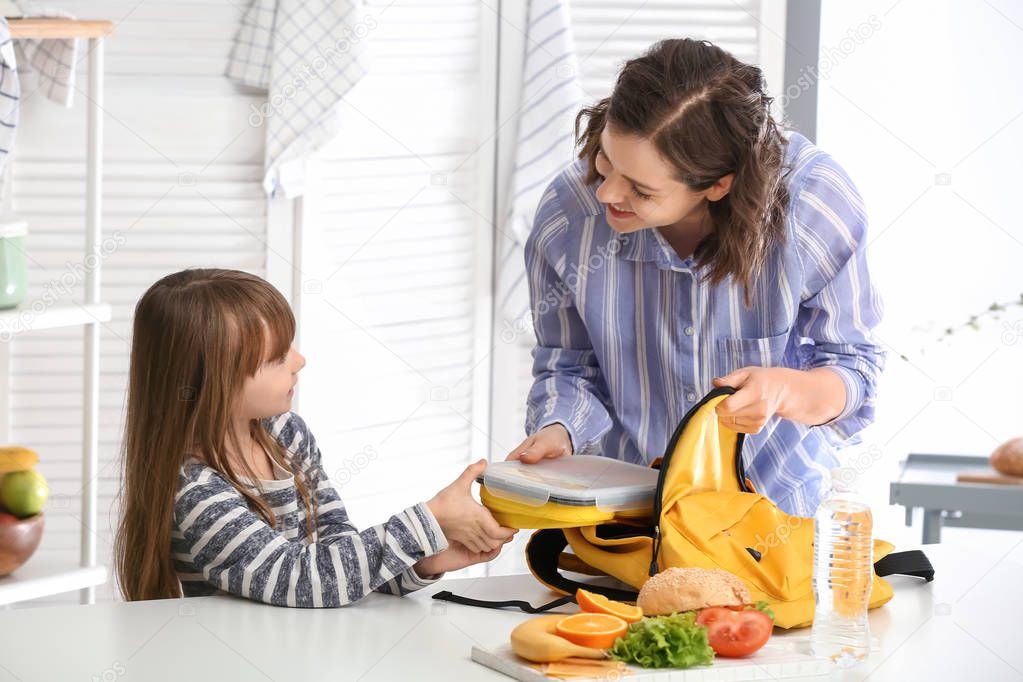 Mother with cute daughter packing lunch box into backpack at home