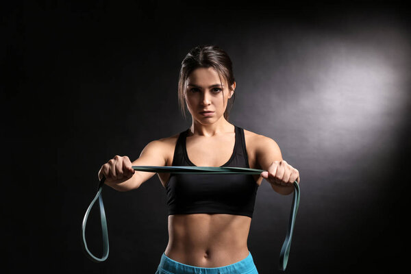 Sporty young woman with resistance band on dark background