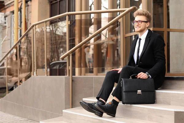 Bonito homem de negócios com pasta elegante sentado em escadas ao ar livre — Fotografia de Stock